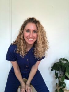 headshot of smiling person leaning forward with curly blonde hair wearing a royal blue jumpsuit