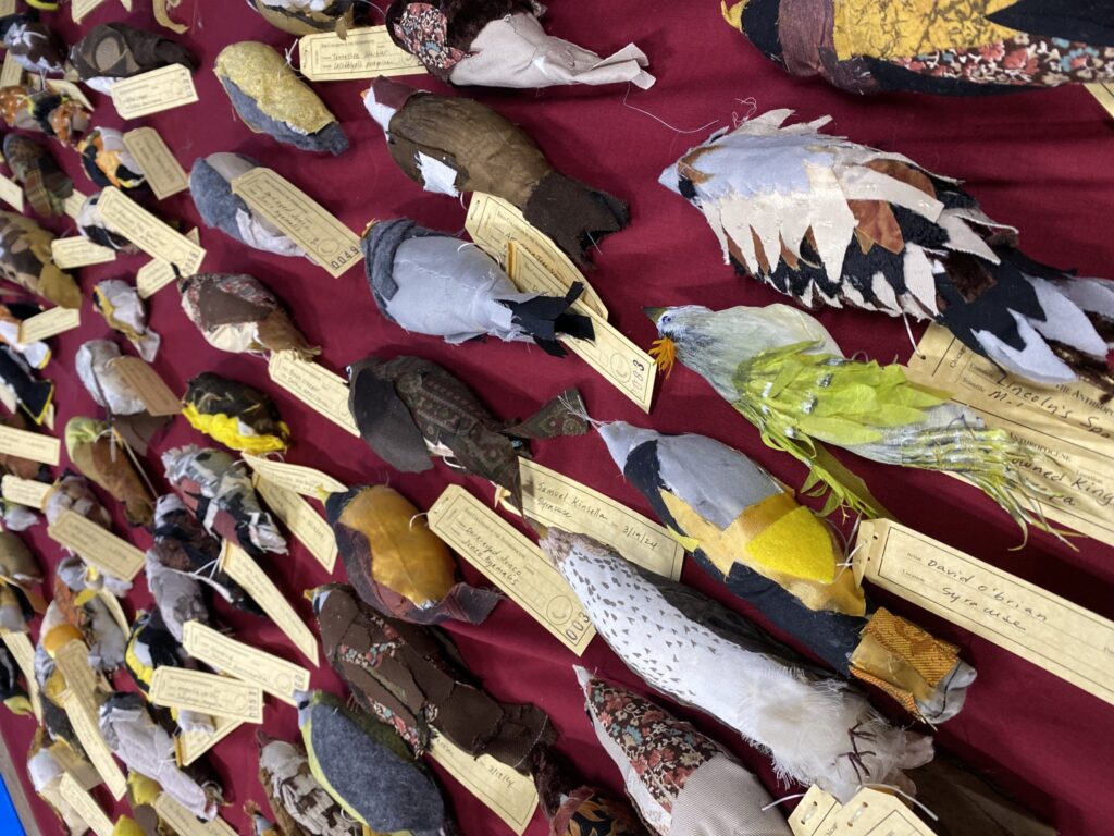 Rows of stuffed birds sewn from colorful scraps of material, each with a tag tied to it with the artist's name, the location, and the species.