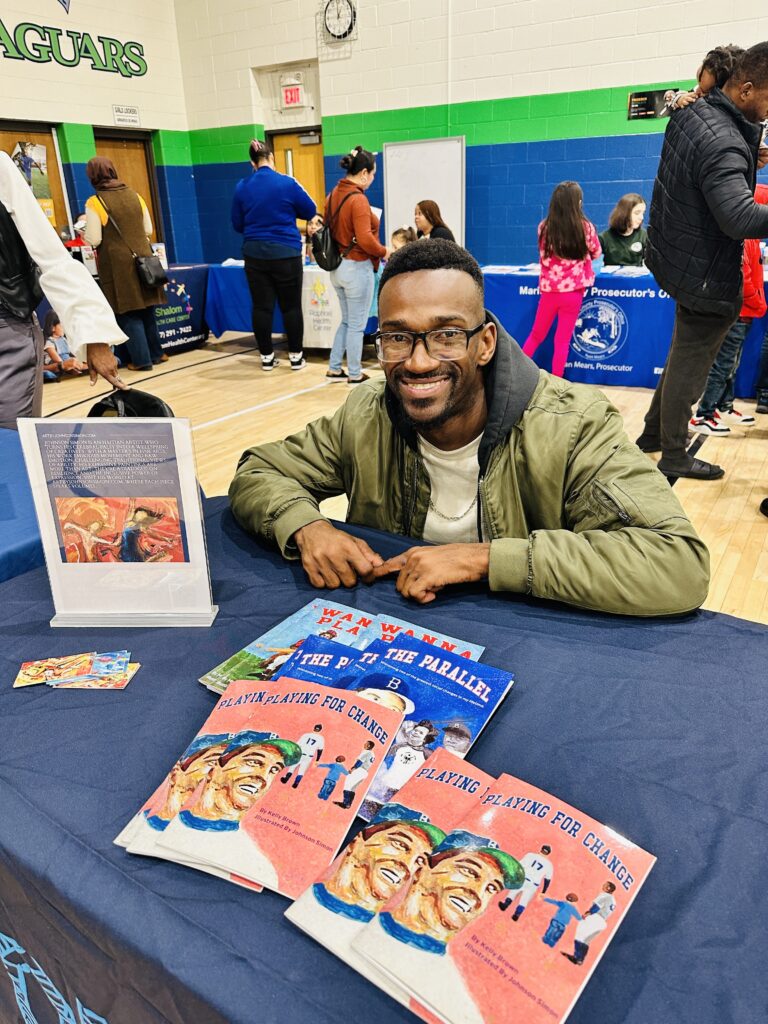 A man sits behind a table at a community event, smiling at the camera. On the table are several books titled Playing for Change along with other illustrated titles. People and display tables are visible in the background.