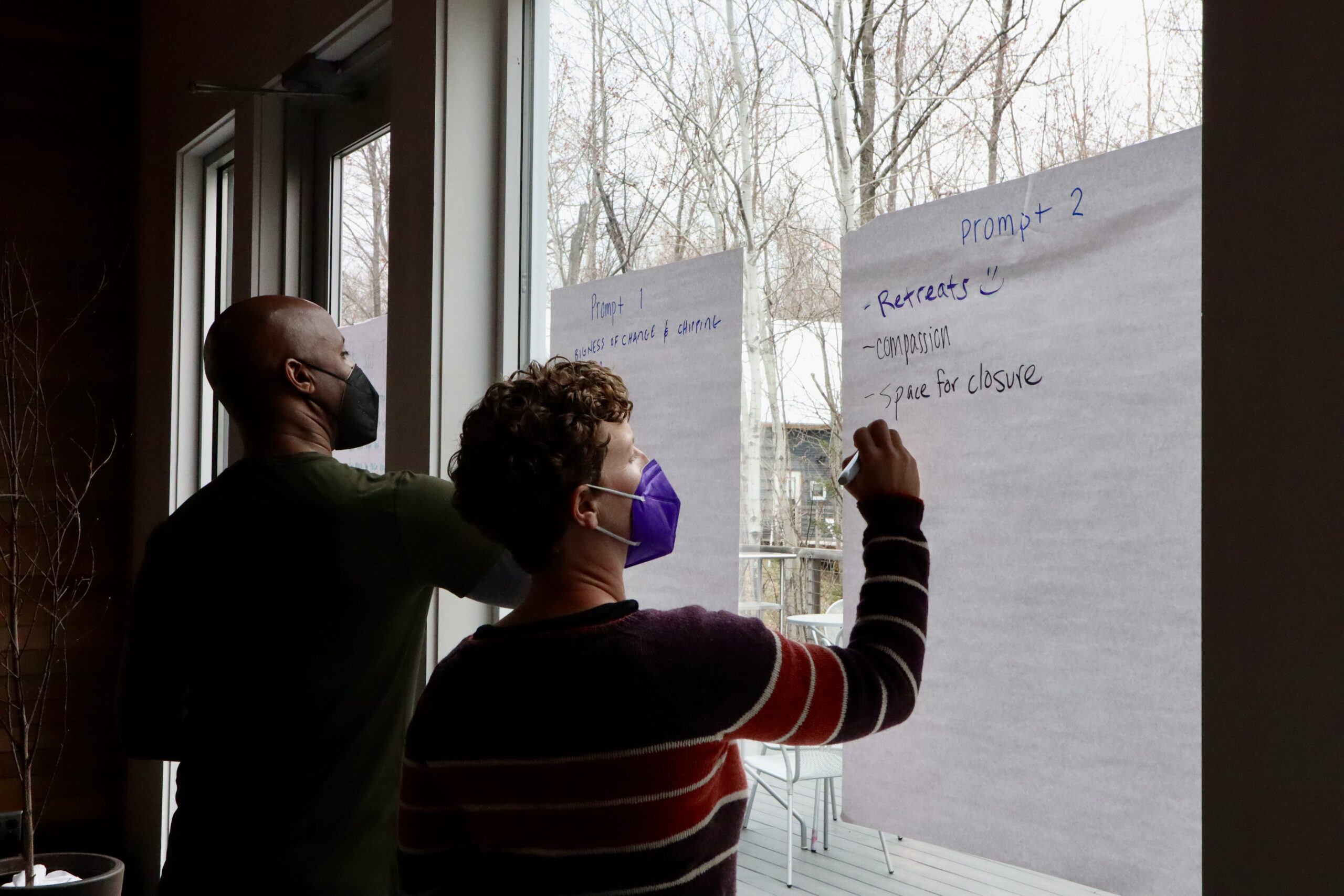 Two people in masks add to large post it notes on a window