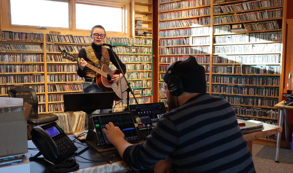 A person with light skin and short hair plays an acoustic guitar in a studio filled with CDs.