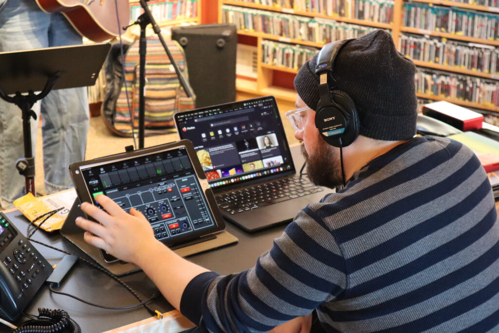 A person with light skin wearing a striped shirt and a beanie and controlling a sound system inside a studio.