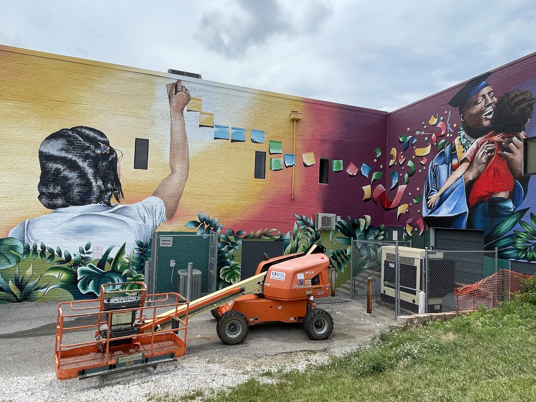 A mural depicting people with dark skin tones. One has their back to the viewer, an arm raised pasting colorful post-it notes. These notes trail to become confetti for a scene depicting a joyful adult in a graduation gown embracing a young child.