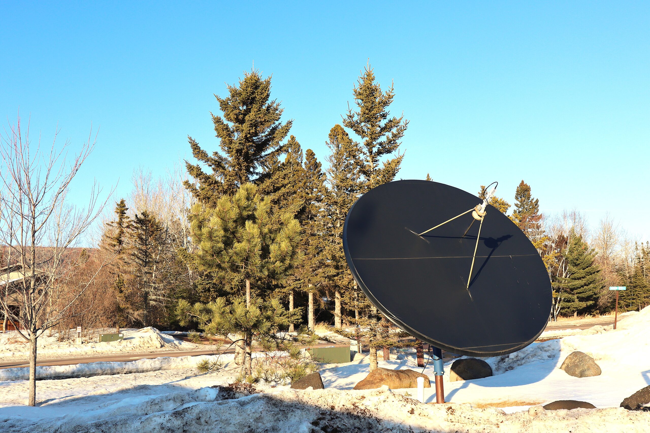 A large radio satellite in the snow next to pine trees and a blue sky.