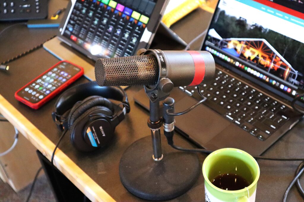 A work desk with an iPad, a laptop, a microphone, a cup of tea, headphones, and a smartphone.