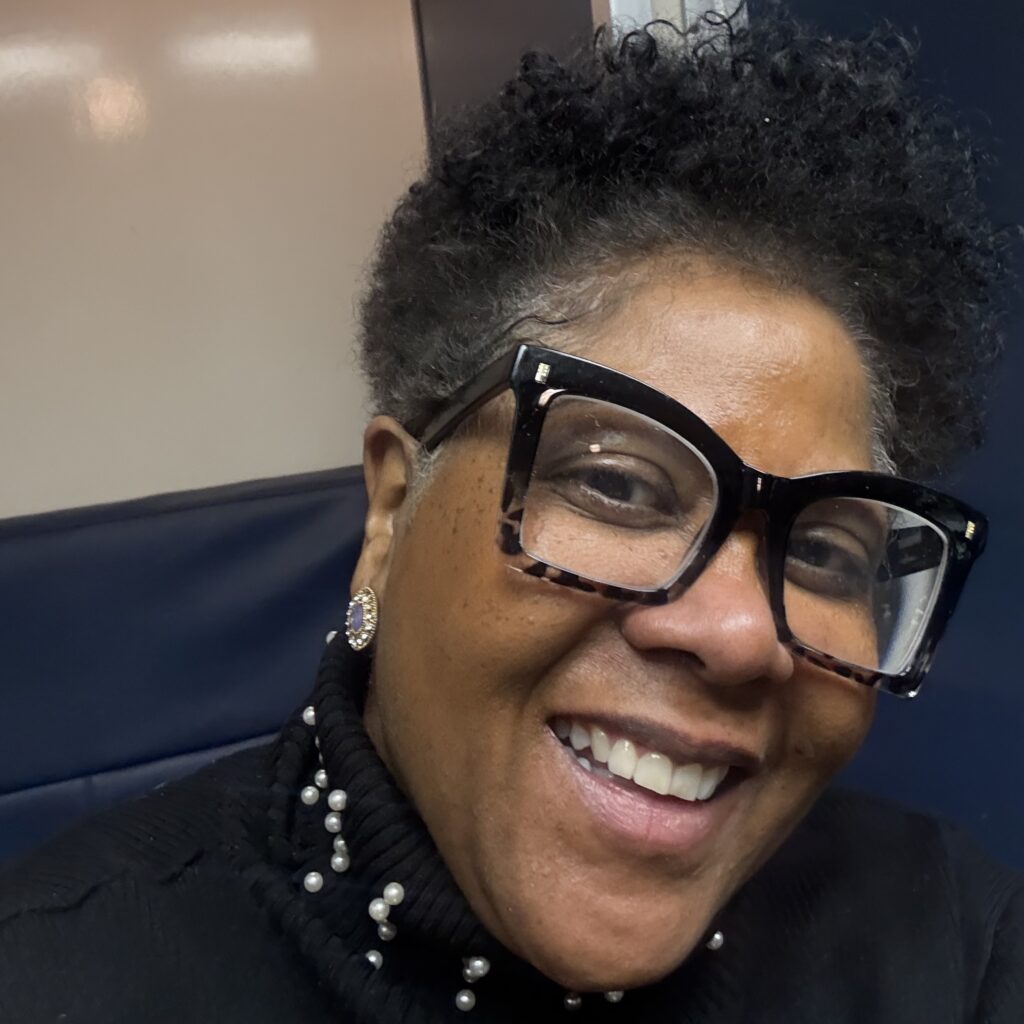 Headshot of a smiling person with medium skin tone, short black hair, and glasses, wearing a black top.