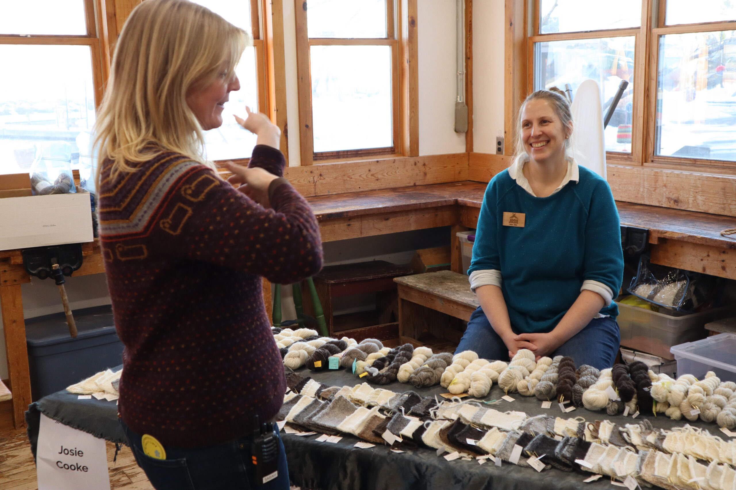 Two women with light skin chatting over a table with yarn and wool samples.