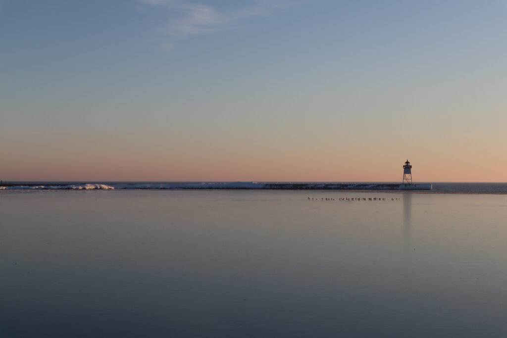A sunset over calm waters, with a lighthouse and a flock of birds.