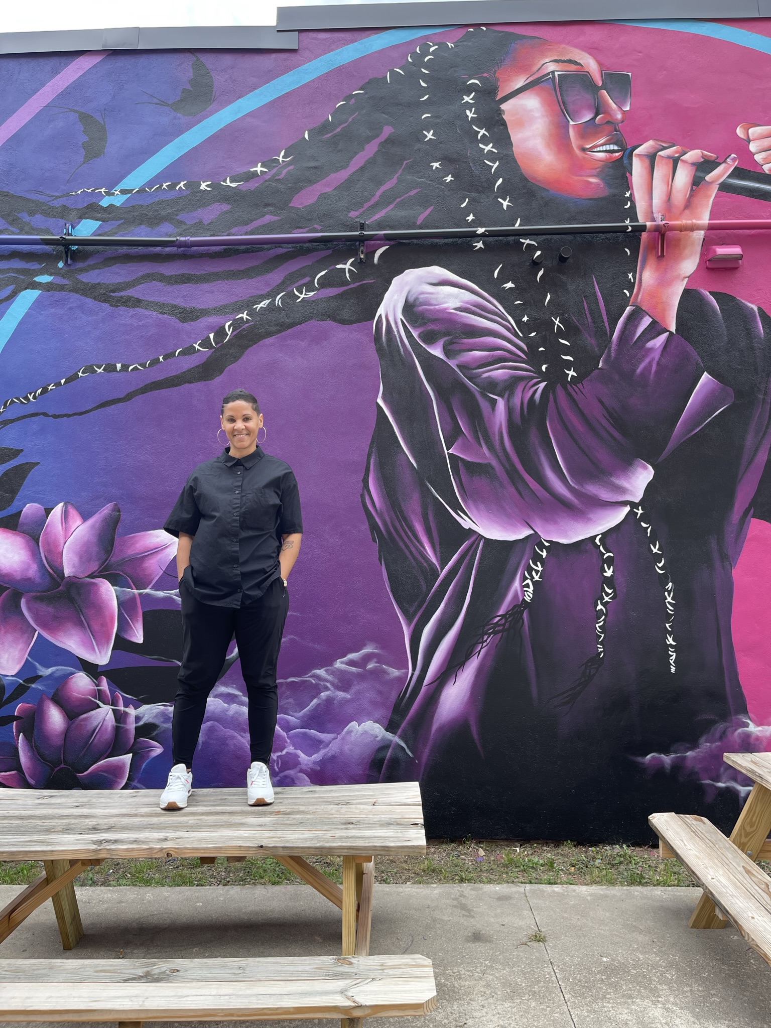 A mural depicting a young Black woman, wearing braids and baby hair, singing into a microphone with one fist in the air. The artist stands in front of the mural.