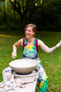 Middle-aged brunette woman in tye-dye overalls sitting at the potters wheel in an outdoor setting with a big smile on her face.
