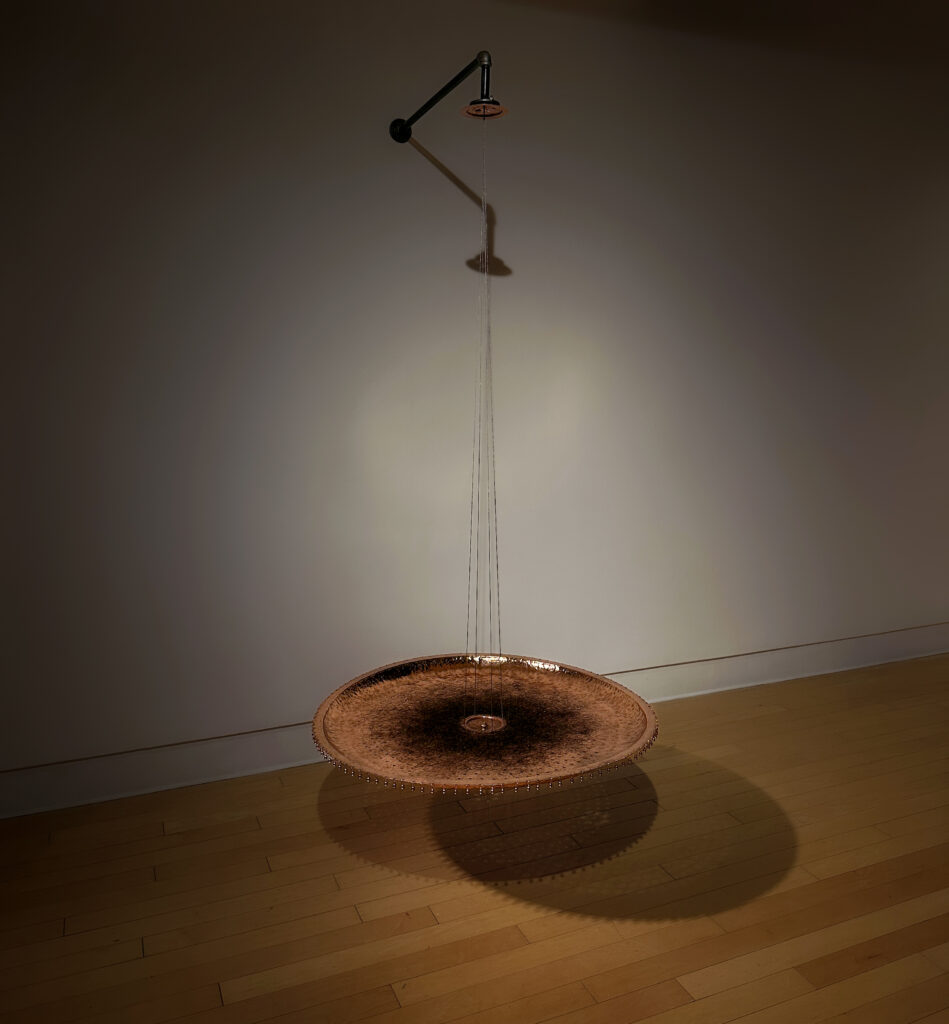 A sculptural installation featuring a copper disc suspended in midair from thin steel wires. Above it, a showerhead drips strands of entangled shed hair down into the center of the disc, forming a dark circular mass. The piece casts a soft shadow onto the wooden floor and wall of the gallery space.
