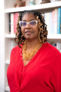Photo of a Black woman with large white glasses and medium-length dredlocks in a bright red dress.. She is standing against a colorful bookshelf.