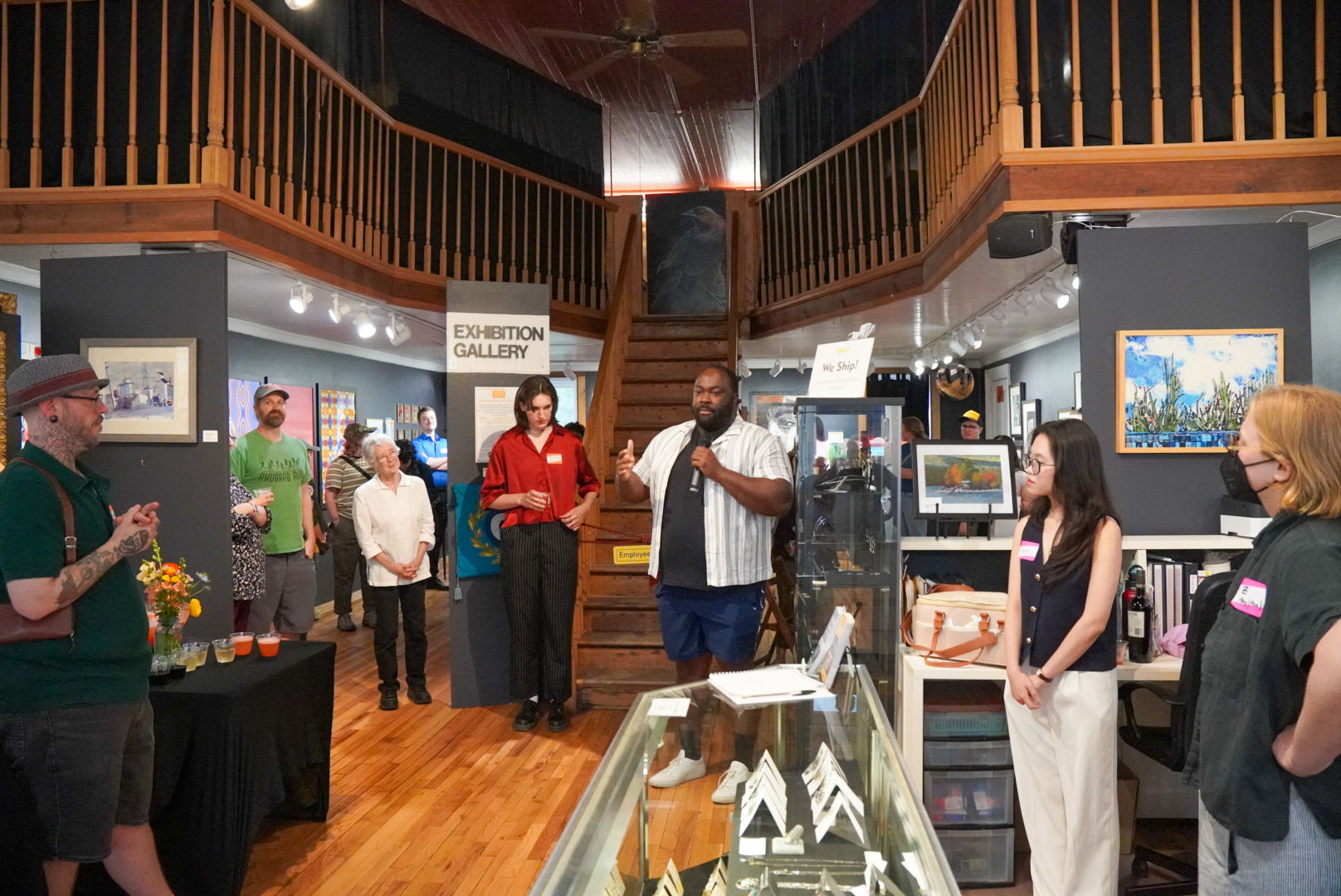 A group of people gather inside the Lanesboro Arts gallery for an event. A speaker holds a microphone while others stand around listening. Artwork hangs on the walls, and a wooden staircase rises to a lofted second floor behind the group.