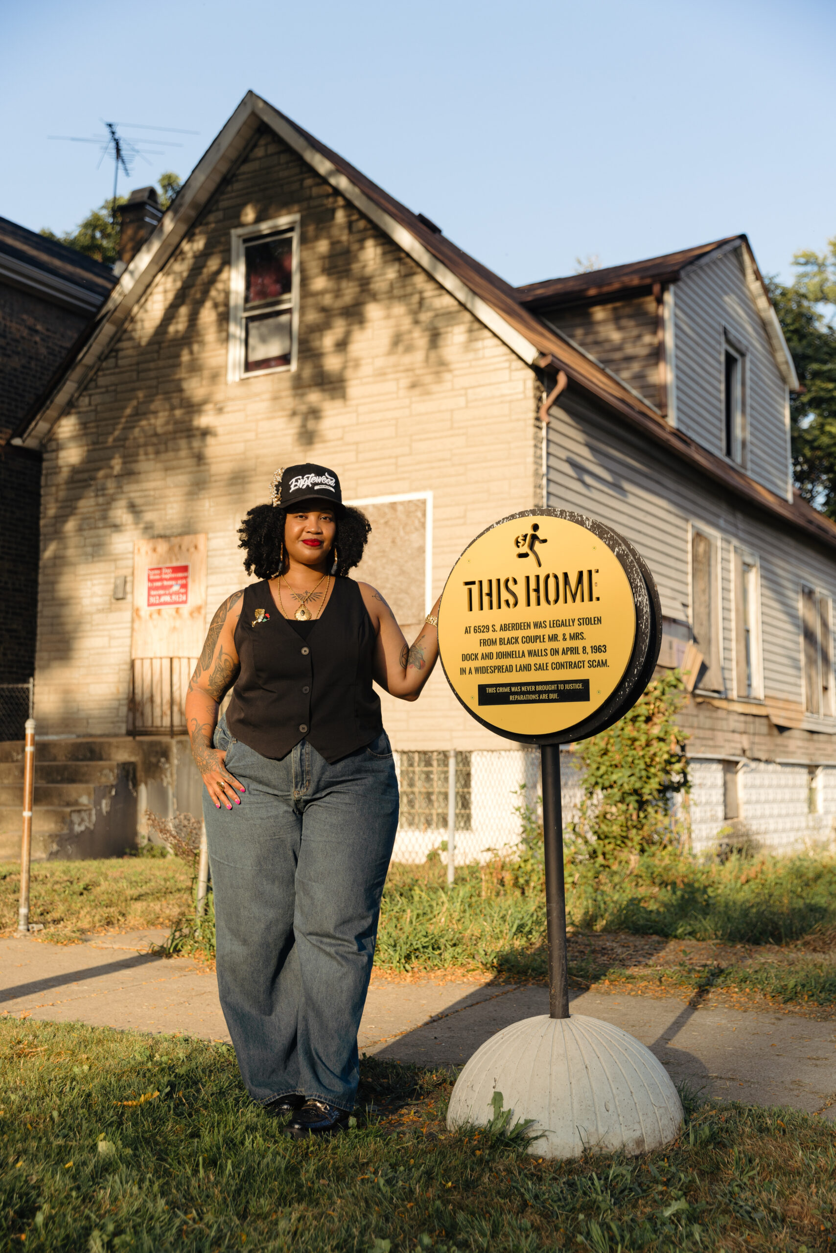 A person stands next to a historical marker sign in front of an old house. The sign reads "This Home at 6529 S. Aberdeen was legally stolen from black couple Mr & Mrs Dock and Johnella Walls on April 8, 1963 in a widespread land sale contract scam. This crime was never brought to justice. Reparations are due."