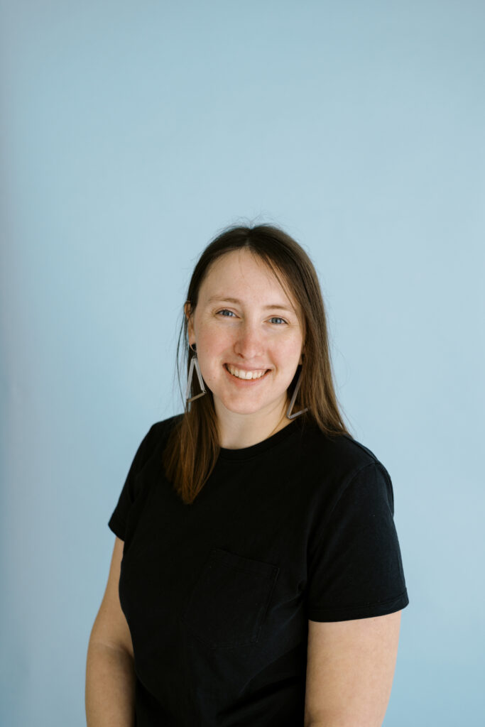 A headshot of a smiling person of light skin tone with straight brown hair wearing a black t-shirt against a light blue background.