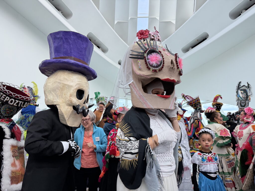 A room full of people, some of whom are in traditional clothing and headgear, imcluding some wearing large paper sculptures over their heads.