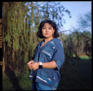 An image of a woman with a neuteral expression, chin length, short curly hair, a tan complexion, in a navy blue loose fitting collard shirt, standing in front of green foliage.