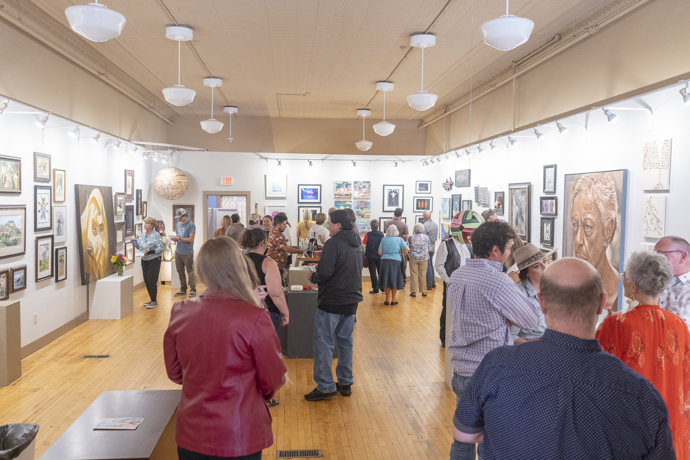 Many people gather in a well lit art gallery, observing artwork on the walls and talking to one another.