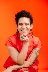 Headshot of a mixed race Black woman, smiling at the camera. Her chin rests in one of her hands, and she is wearing all orange with an orange background. She has short curly black hair and is wearing jewelry.