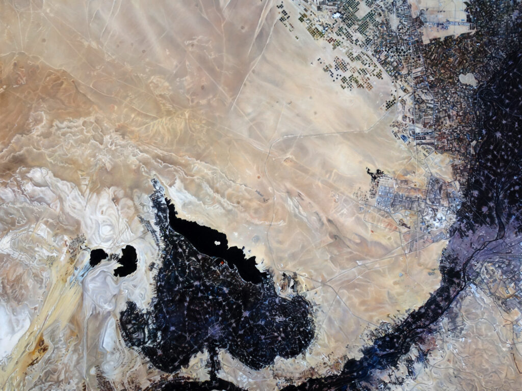 An abstract aerial view of a pale sandy desert with swirls of white, black bodies of water, and tiny human settlements that include roads and buildings.