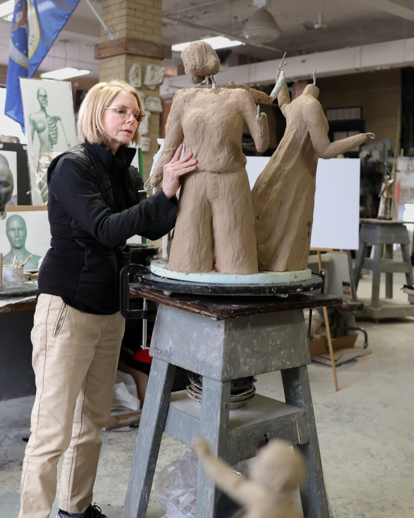 An artist shapes a clay sculpture of two women figures in a studio, working on the early stages of the Ohio Women’s Monument design.