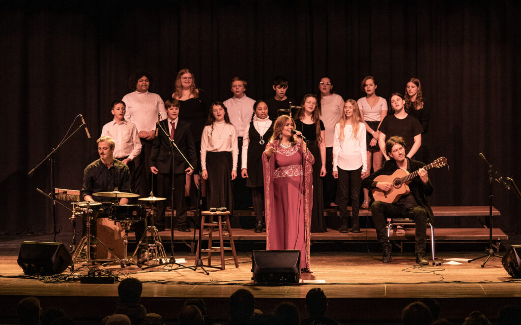 A vocalist in a colorful Jordanian robe flanked by a percussionist, guitarist, and choir of middle schoolers.