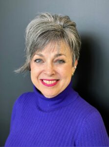 A woman with light skin and short, styled salt-and-pepper hair, wearing a purple turtleneck and smiling.