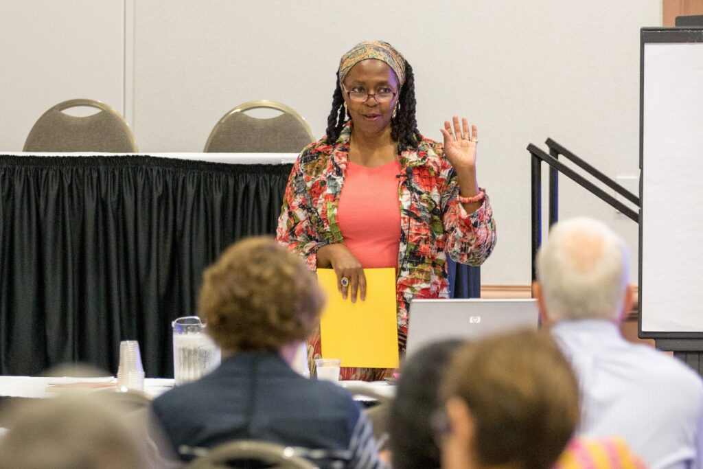 A woman stands at the front of a conference hall speaking to a seated crowd during a session.