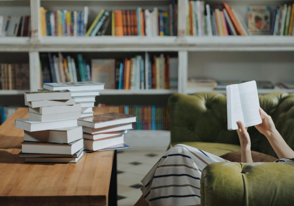 A person in striped skirt reads a book on a green sofa, surrounded by a wooden table stacked with books. Shelves filled with books line the background.