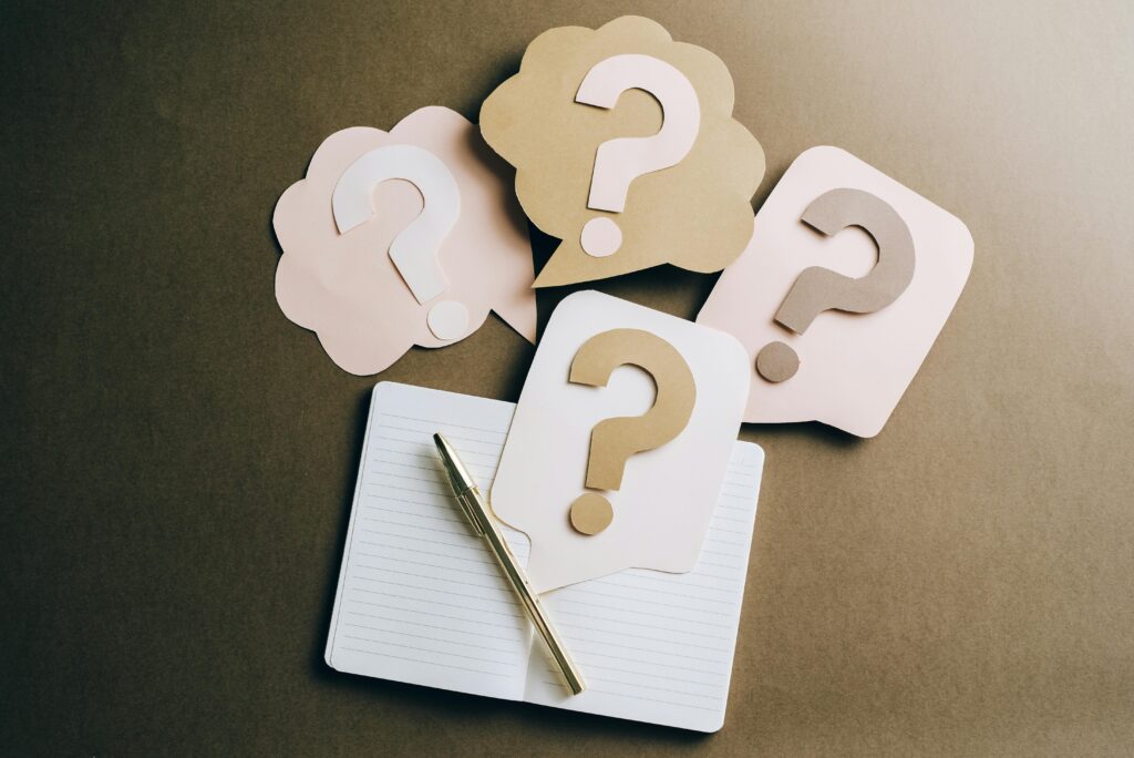 A notebook open to a blank page with a gold pen and four question marks made out of cut paper.