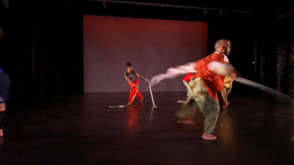 Two dancers in red costumes perform on a darkened stage, sweeping long white ribbons through the air against a red-lit backdrop, their motion captured mid-blur conveying energy and speed.