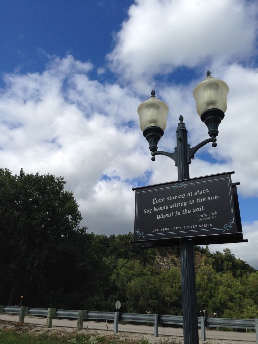 A black streetlamp in Lanesboro, Minnesota holds a poetry sign that reads, “Corn staring at stars. Soy beans sitting in the sun. Wheat in the soil.” Trees and a bright blue sky with clouds fill the background.