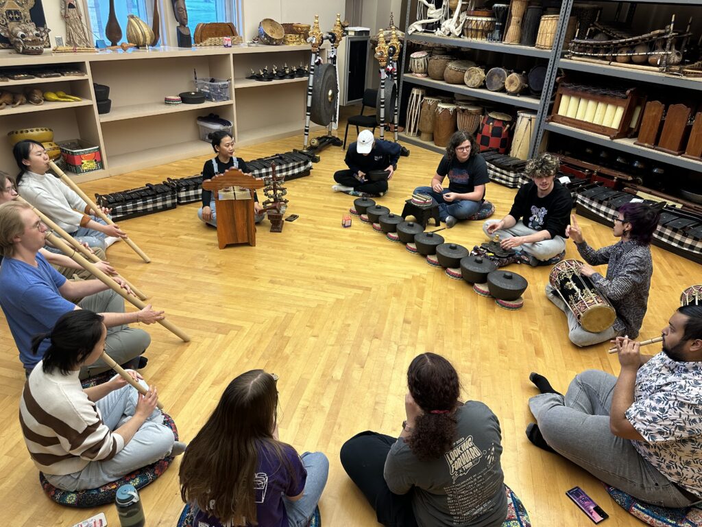 A group of people sitting in a circle surrounded by shelves that hold different kinds of musical instruments. Most of them are in the process of playing an instrument.