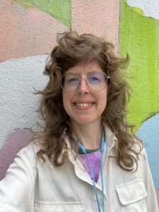 Headshot of a smiling person with glasses, light skin tone and long layered brown hair, wearing layered shirts under a white long sleeve, blue headphones around her neck, standing in front of a colorful wall