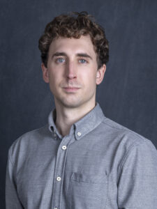 Headshot of a person smiling, with light skin tone, short curly brown hair, with blue eyes, and a grey button up collared shirt.