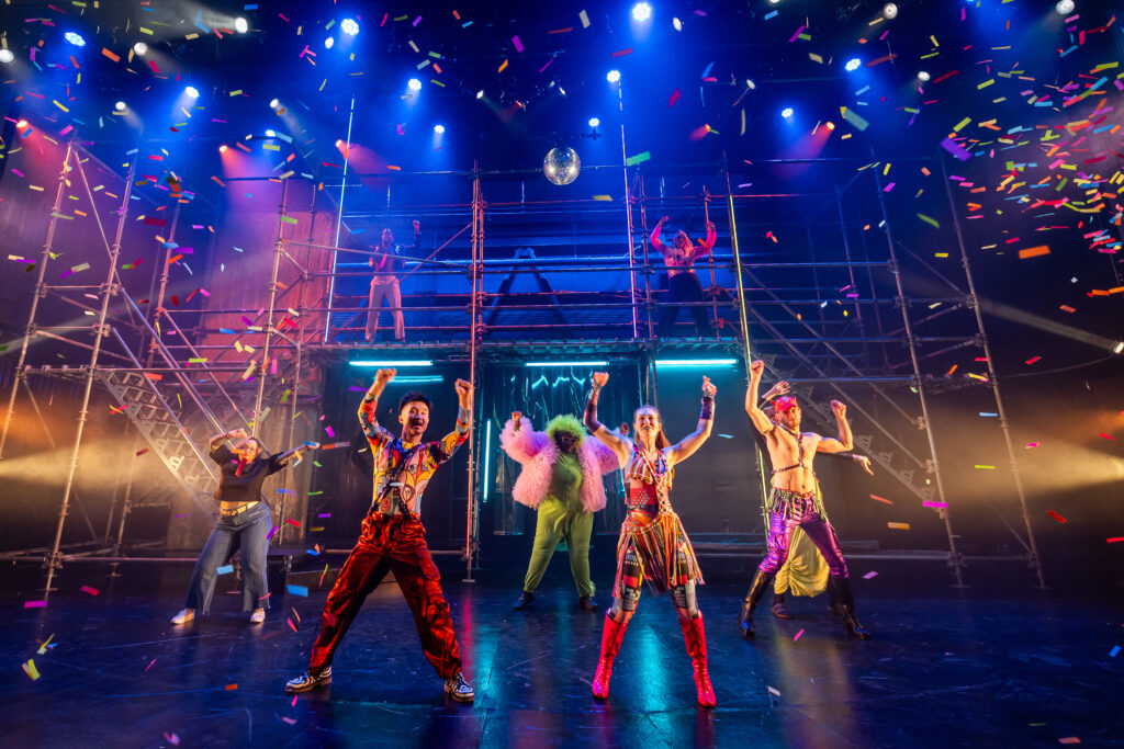 Five actors in brightly colored costumes standing below a silver disco ball with their arms in the air on a black stage while colorful confetti showers down from the ceiling. Two more actors stand on a raised platform behind them supported by metal scaffolding.