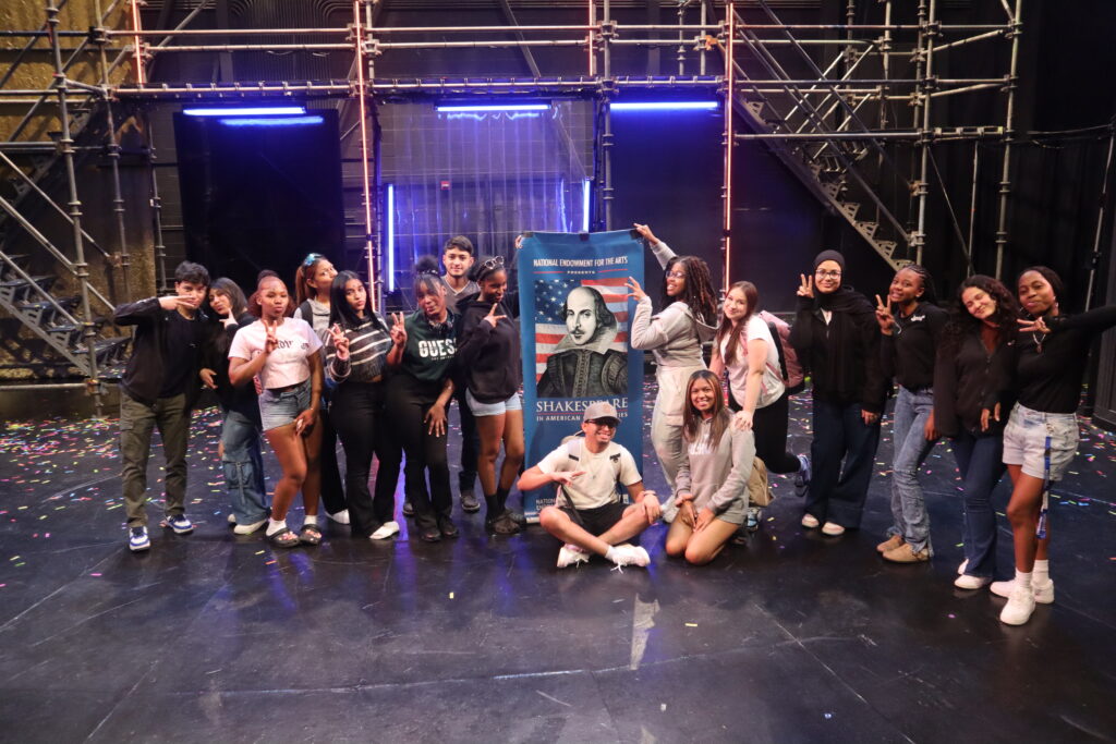 Sixteen teenaged students standing on a black stage in front of metal scaffolding, posing next to a blue banner with an image of William Shakespeare superimposed over an American flag entitled "National Endowment for the Arts Presents Shakespeare in American Communities."