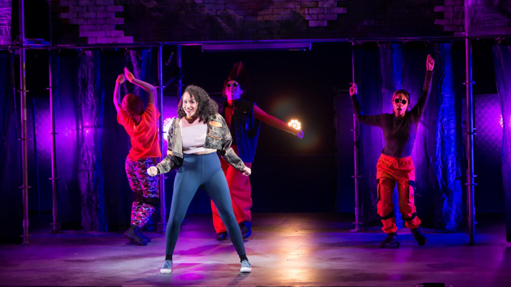 Four people dancing on a stage cast in a dim purple light; the person in front has a spotlight on them and they're smiling at the camera. Two people in sunglasses dance behind them, and another person dances with their back to the camera.