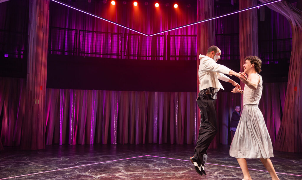 A tall light skinned person wearing a white skirt stands to the left, holding hands with a person jumping in the air. There is a purple curtain in the background.