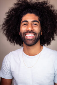 Headshot of a smiling man with brown skin tone and big curly hair, wearing a light blue shirt.