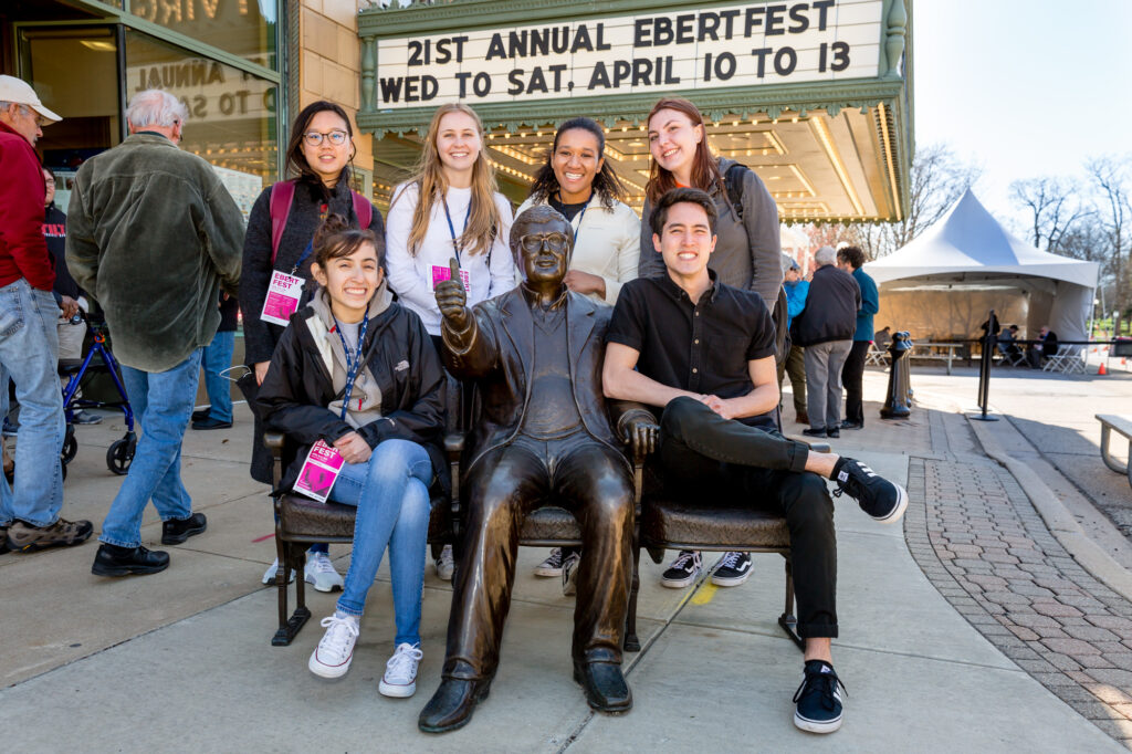 A group of young people smiling and posing next to a bronze statue of a person sitting on a bench gesturing with a thumbs up.