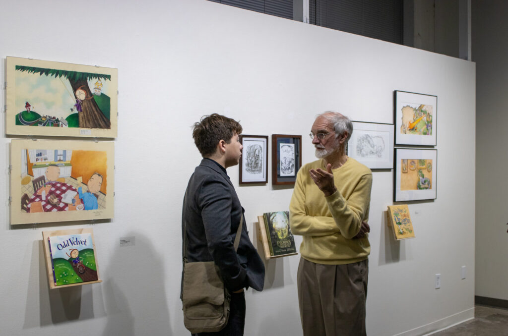 An older adult in a yellow sweater and glasses talking to a younger person with short brown hair and a gray blazer inside an art gallery. On the walls behind them are illustrations from children's books, with the books displayed below the framed artwork.