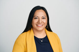 Headshot of a smiling person with light brown tone and long dark brown hair, wearing a yellow jacket and blue shirt, and gold hoop earrings.