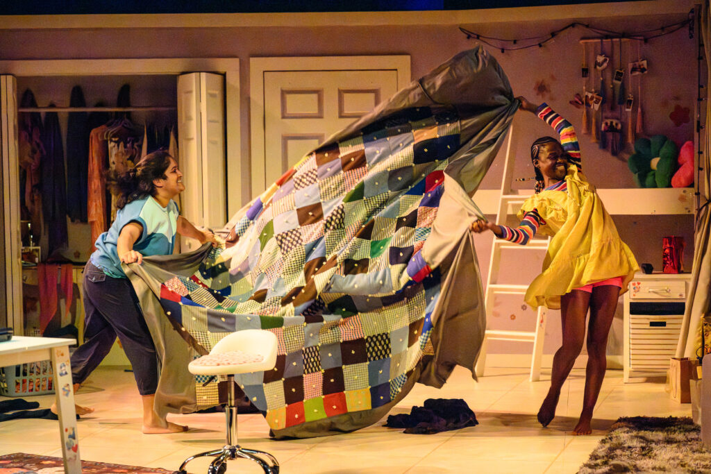 Two performers on a warmly lit stage laugh as they shake out a large patchwork quilt between them. The set depicts a cozy bedroom with clothes, string lights, and wall decorations.