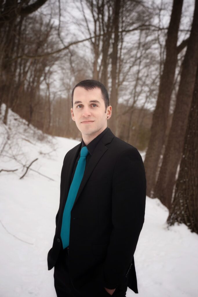 A person of light skin tone wearing a black suit and a teal necktie stands outdoors. There is snow and trees in the background.