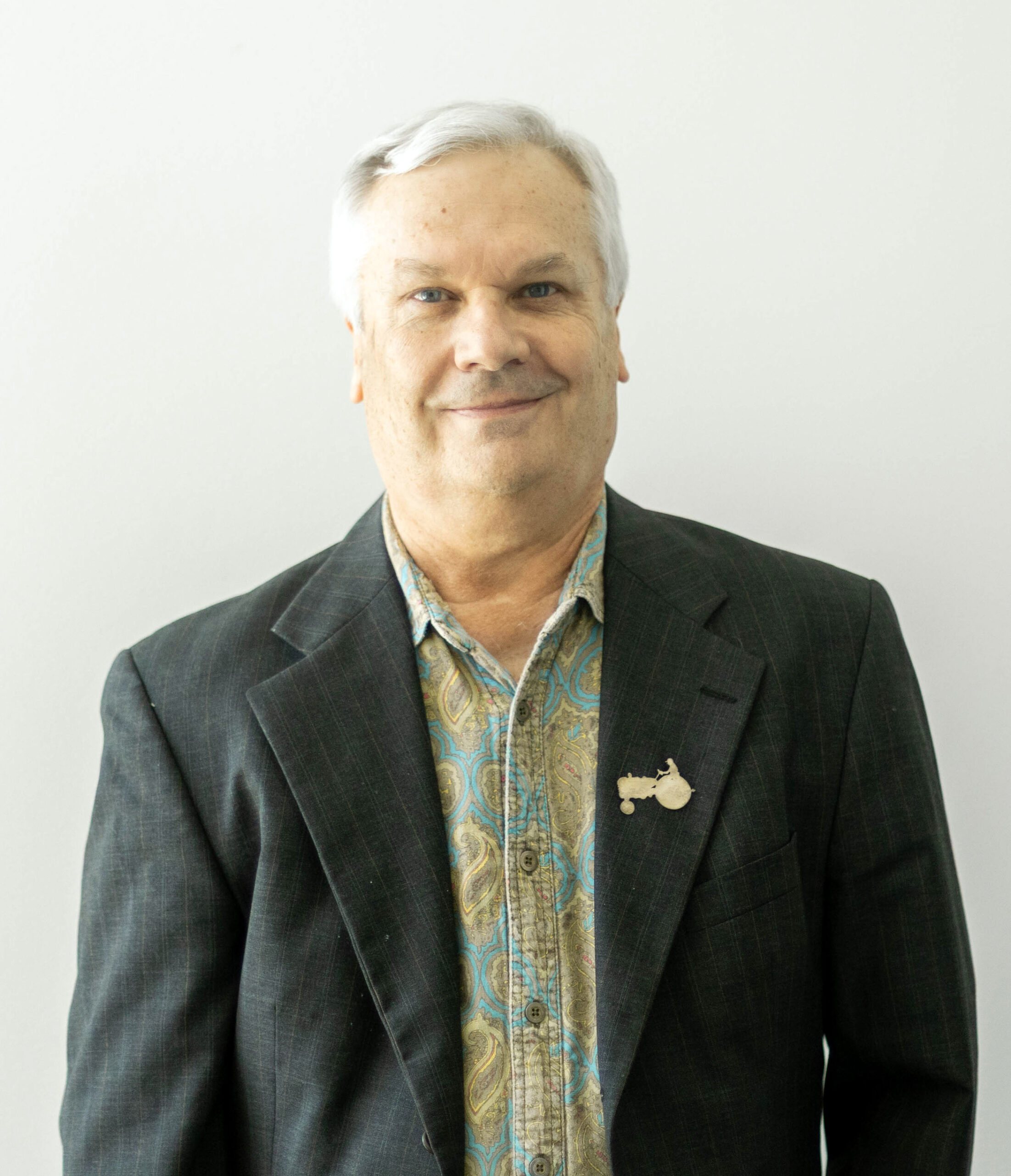 A portrait of John Davis standing against a plain white wall. He wears a dark blazer over a paisley shirt and smiles softly at the camera. A small metal tractor pin is attached to his lapel.