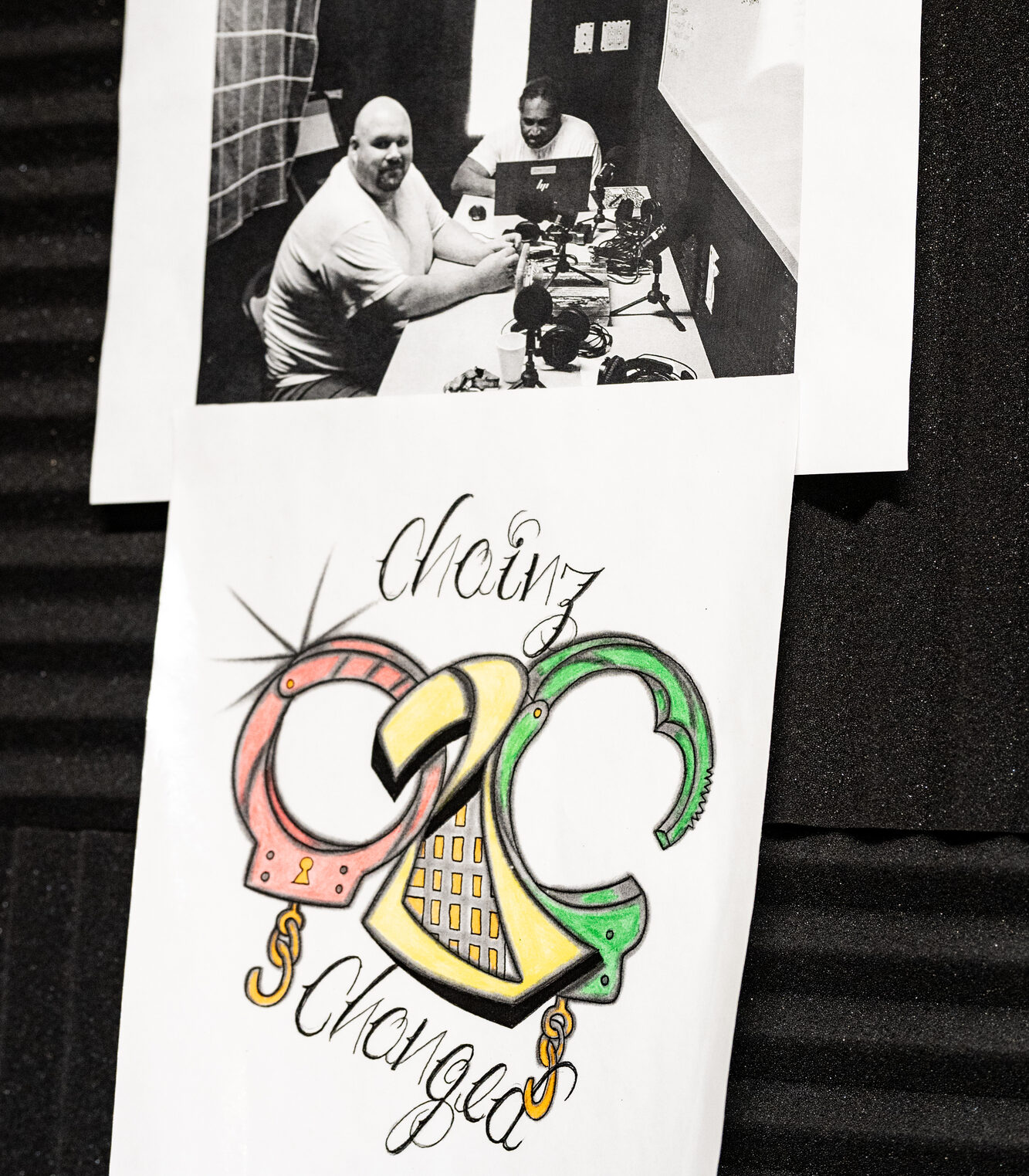 Two sheets of paper stuck on a dark wall. One is a photograph of two people sitting at a table with audio recording equipment. The other is a drawing depicting handcuffs with decorative text that reads "Chainz 2 Changed"