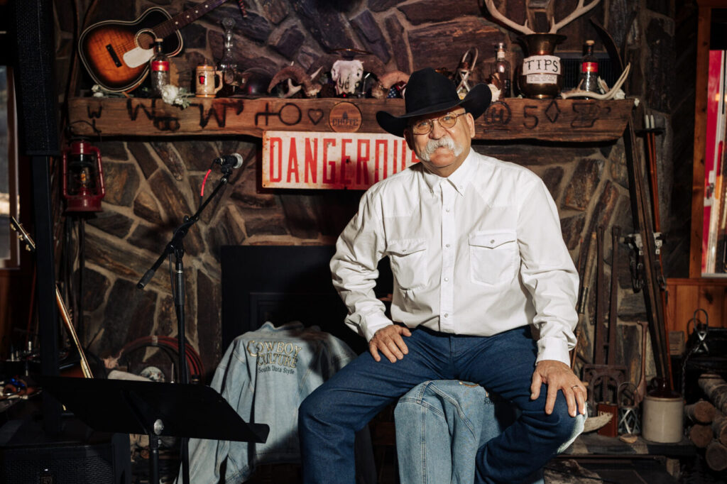 A person with a white mustache sitting on a tall stool and smiling. They are wearing a cowboy hat, thin-framed glasses, a white button-up shirt, and blue jeans.