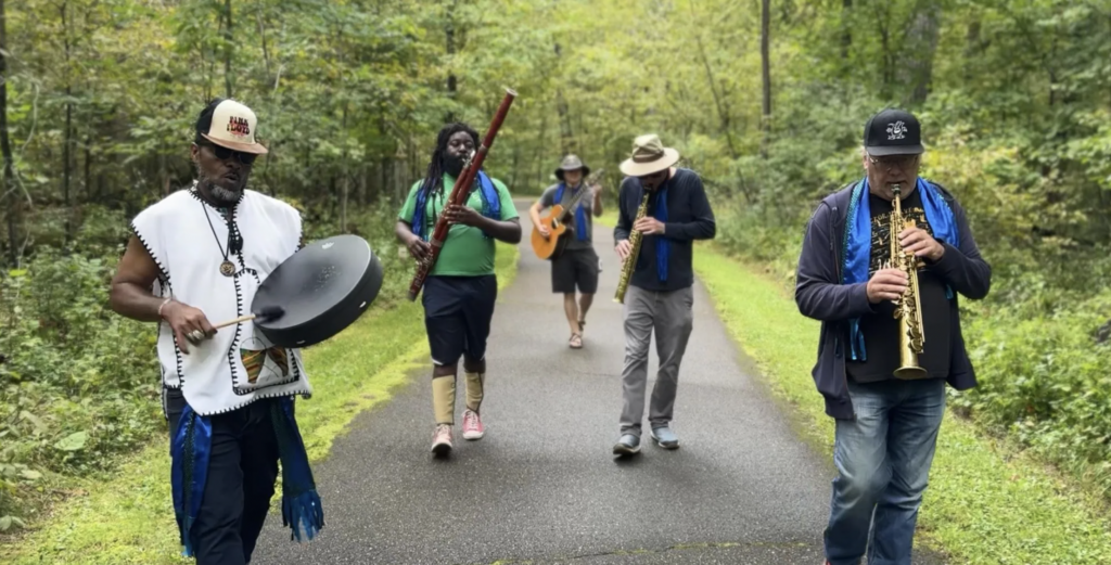 Four musicians walk along a tree-lined path playing a hand drum, bassoon, acoustic guitar, and soprano saxophone. Lush green forest surrounds them.