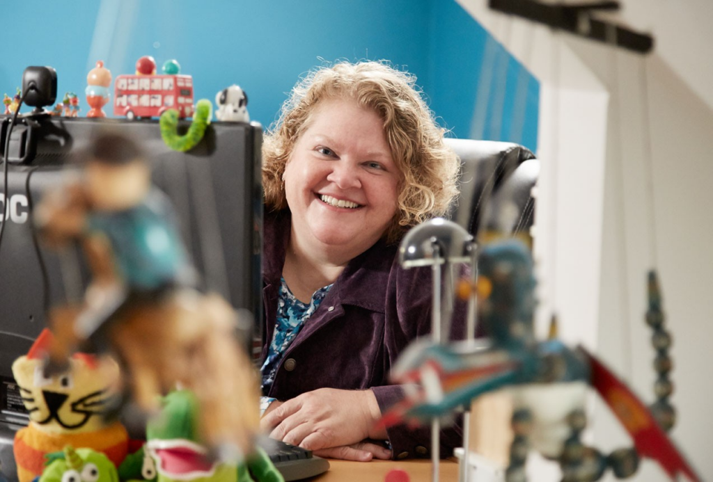 A light-skinned woman smiling at the camera sitting at a crowded desk full of toys.